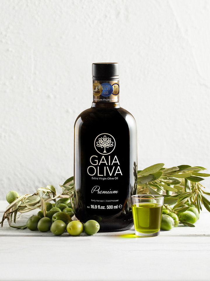 Bottle of Ayvalik Early Harvest Extra Virgin Olive Oil with green olives and glass of olive oil on a white background