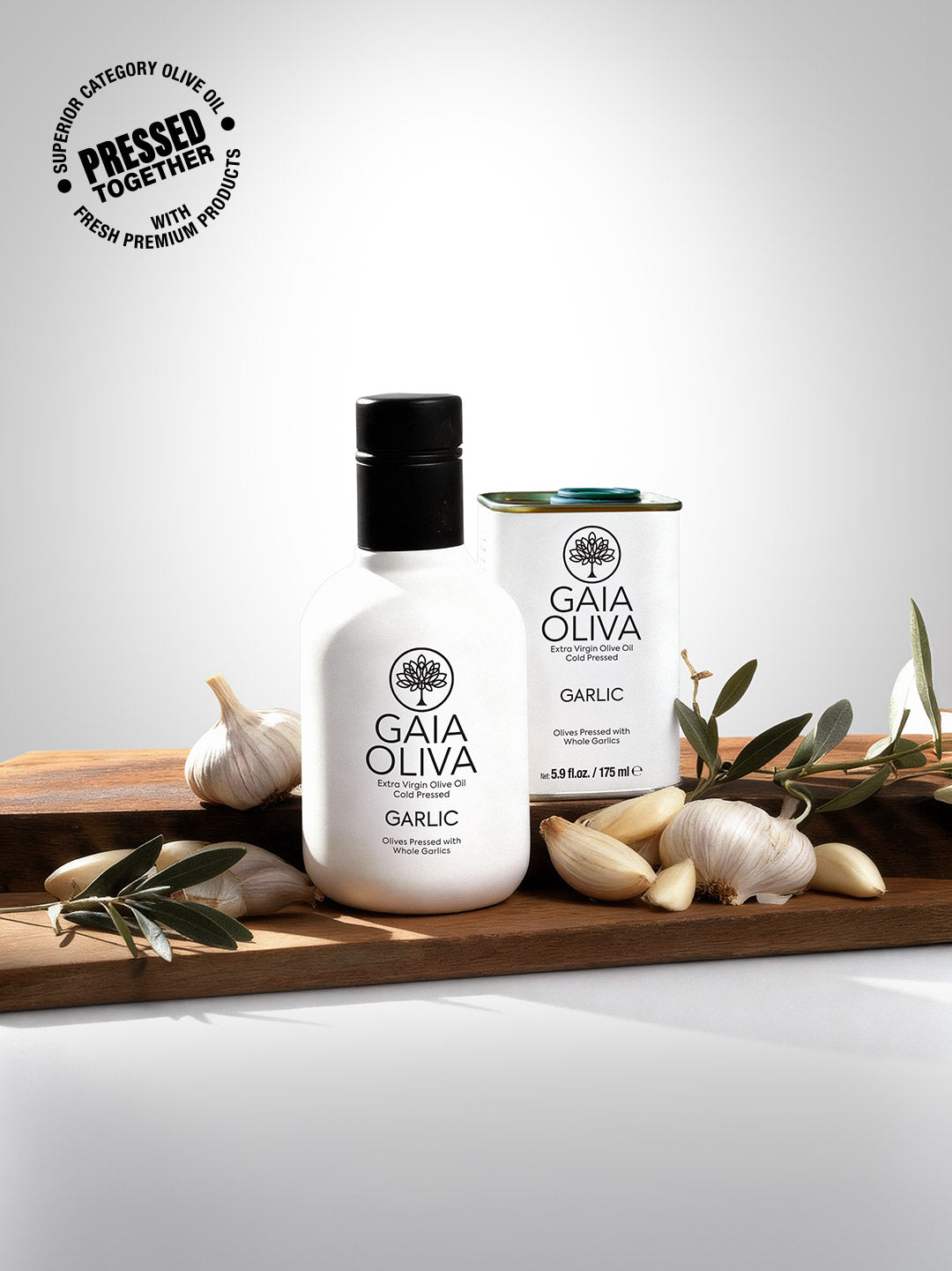 Bottle of Gaia Oliva Garlic Extra Virgin Olive Oil with fresh garlic cloves and smal tin on wooden surface.