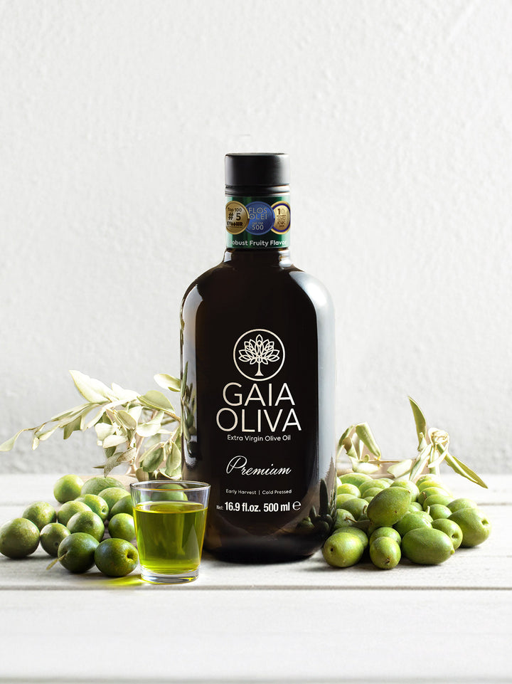 Bottle of Memecik Early Harvest Extra Virgin Olive Oil with a glass of olive oil and green olives on a white surface.